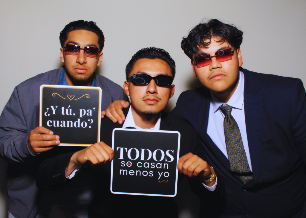 Three young men in suits posing at a photo booth with funny Spanish wedding signs